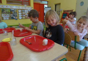 Dzieci siedzi przy stoliku, dziewczynka obserwuje reakcje wody utlenionej z drożdżami w swoich pojemnikach. Chłopiec miesza składniki za pomocą łyżeczki.